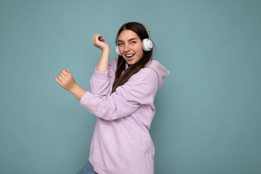 Attractive positive smiling young brunette woman wearing light purple hoodie isolated over blue background wall wearing white bluetooth headphones listening to good music and having fun looking at camera.