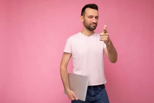 Handsome brunet man holding laptop computer and pointing on you looking at camera in t-shirt on isolated pink background.