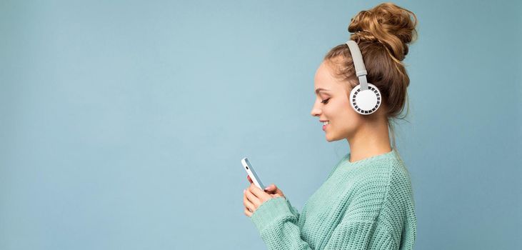 Panoramic Side-profile photo shot of beautiful joyful smiling young female person wearing stylish casual outfit isolated over colorful background wall wearing white bluetooth wireless earphones and listening to music and using mobile phone looking at gadjet display. copy space