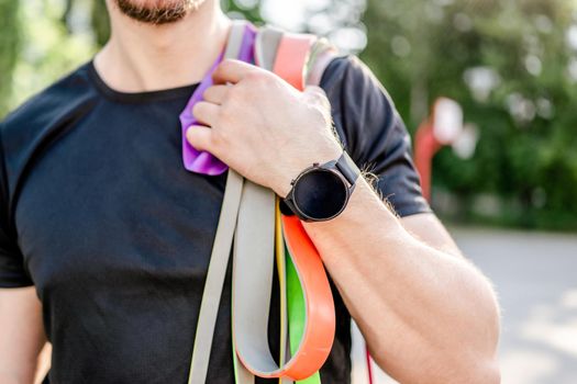 Sportive man guy holding in his hand wearing fitness tracker set of colorful elastic rubber bands at the stadium outdoors. Athlete male person during workout with additional sport equipmant