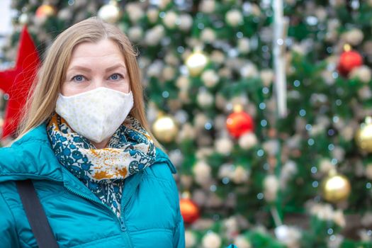 A woman in a protective mask near a festively decorated Christmas tree on the street. Christmas and New Year during the coronavirus pandemic.