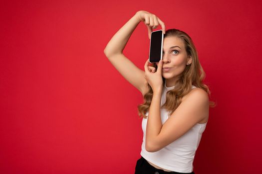 Shot of pretty funny young blonde female person wearing white t-shirt isolated on red background with copy space holding smartphone showing phone in hand with empty screen for cutout looking up to the side.