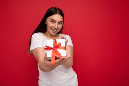 Beautiful happy young brunette woman isolated over colourful background wall wearing stylish casual clothes holding gift box and looking at camera. Copy space, mockup