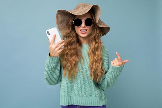 Beautiful smiling young blonde woman wearing sunglasses and hat isolated on background with copy space holding smartphone looking at camera.