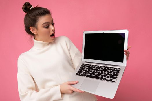 Photo of beautiful amazed and surprised woman with gathered brunette hair wearing white sweater holding computer laptop and looking at the open netbook isolated over pink wall background.