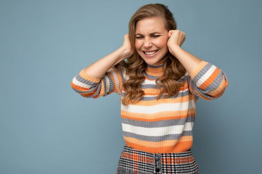 Photo of emotional young attractive blonde curly woman with sincere emotions wearing casual striped pullover isolated on blue background with copy space and covering ears trying not to hear.