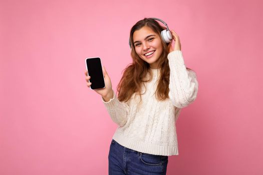 Attractive positive smiling young woman wearing stylish casual outfit isolated on colourful background wall holding and showing mobile phone with empty screen for cutout wearing white bluetooth headphones and having fun looking at camera. copy space