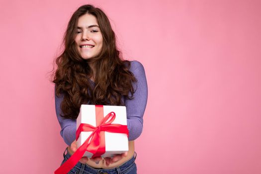 Shot of attractive positive smiling young brunette woman isolated over colourful background wall wearing everyday trendy outfit holding gift box and looking at camera. Copy space