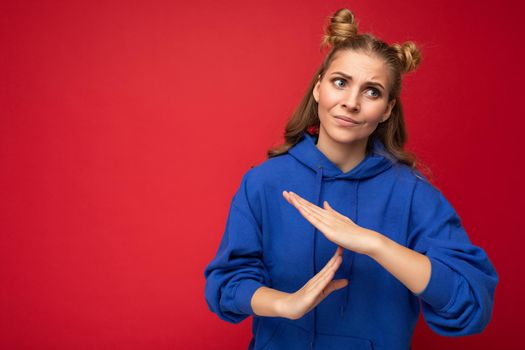 Photo of emotional adult attractive blonde woman with two horns with sincere emotions wearing hipster bright blue hoodie isolated over red background with copy space and showing timeout sign.