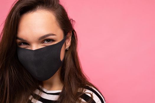 Woman wearing stylish protective face mask, posing on pink background. Trendy fashion accessory during quarantine of coronavirus pandemic. Close up studio portrait. Copy, empty space for text.