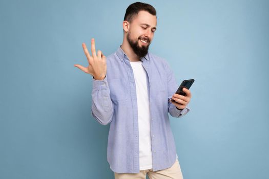 Photo shot of handsome happy smiled satisfued positive good looking young man wearing casual stylish outfit poising isolated on background with empty space holding in hand and using mobile phone messaging sms looking at smartphone display screen.