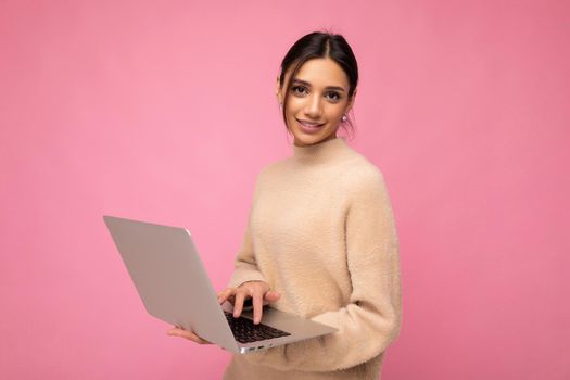 Photo of Beautiful young brunette woman wearing beige sweater holding netbook computer typing text on keyboard looking at camera isolated over pink background.