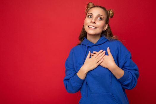 Portrait of young happy positive beautiful blonde woman with two horns with sincere emotions wearing casual bright blue hoodie isolated on red background with empty space and holding hands on heart.