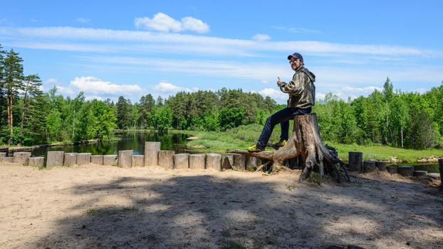 Smiling man tourist is sitting on a tree stump side of the river.Summer forest