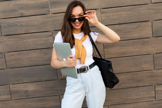 Photo of beautiful fascinating charming smiling young brunette girl wearing white t-shirt light blue jeans and yellow sweater holding computer laptop and black sunglasses staying at the street and looking at camera.