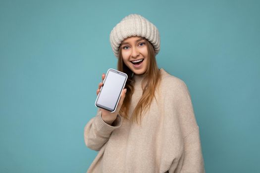 Photo of beautiful positive joyful young blond woman wearing stylish beige sweater and beige knitted winter hat poising isolated over blue background with empty space holding in hand and showing mobile phone with empty screen for mockup looking at camera.