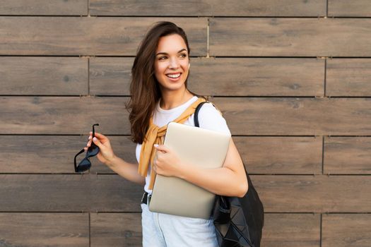 Close-up portrait of nice-looking charming attractive lovely fascinating cheerful cheery straight-haired brunet woman looking to side in the street with laptop computer and glasses in hands