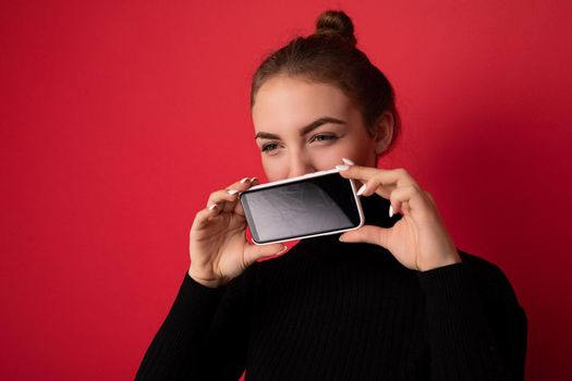 Closeup portrait Photo of beautiful young brunette woman wearing black sweater standing isolated over red background showing mobile phone with empty screen for mockup looking to the side.