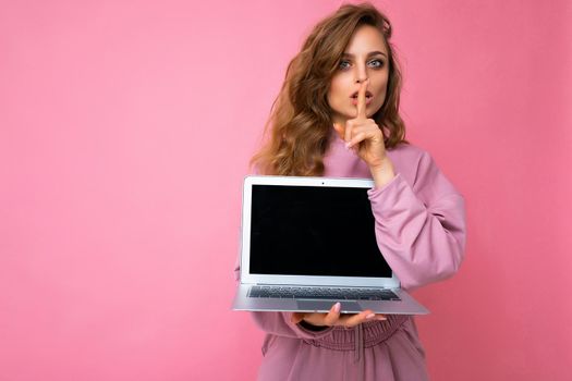 Close-up portrait of beautiful dark blond curly young woman holding laptop computer looking at camera holding finger close to mouth and lips showing gesture shhh and saying hush be quiet wearing pink sweater isolated over pink wall background. Mock up copy empty space.
