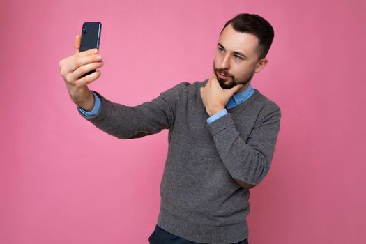 Photo of positive handsome young brunette unshaven man with beard wearing casual grey sweater and blue shirt isolated on pink background wall holding smartphone taking selfie photo looking at mobile phone screen display and thinking.