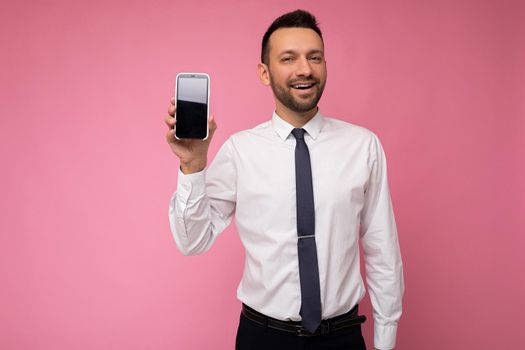 Photo of handsome good looking man wearing casual white shirt and tie isolated on pink background with empty space holding in hand and showing mobile phone with empty screen for mockup looking at camera.