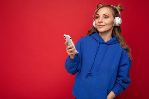 Photo of beautiful joyful smiling young woman wearing stylish casual clothes isolated over background wall holding and using mobile phone wearing white bluetooth headphones listening to music and having fun looking to the side.