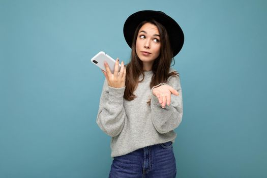 young asking dissatisfied brunette woman pointing hand at you wearing black hat and grey sweater holding smartphone looking to the side isolated on background.