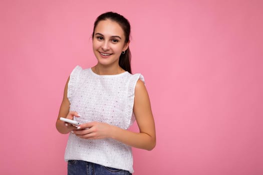 Attractive positive girl wearing white blouse isolated over pink background wall using mobile phone surfing on the internet online looking at camera. Copy space