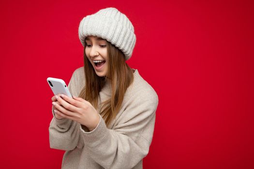 Beautiful amazed young blonde woman wearing casual beige sweater and beige hat isolated over red background holding in hand and using mobile phone communicating online on the internet looking at gadjet display.