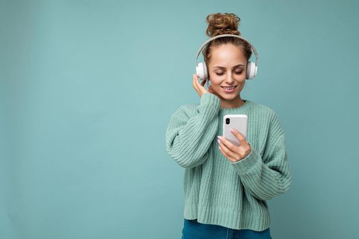 Photo shot of beautiful joyful smiling young female person wearing stylish casual outfit isolated over colorful background wall wearing white bluetooth wireless earphones and listening to music and using mobile phone looking at gadjet display