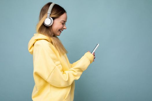 Side-profile photo shot of beautiful joyful smiling young female person wearing stylish casual outfit isolated over colorful background wall wearing white bluetooth wireless earphones and listening to music and using mobile phone looking at gadjet display.