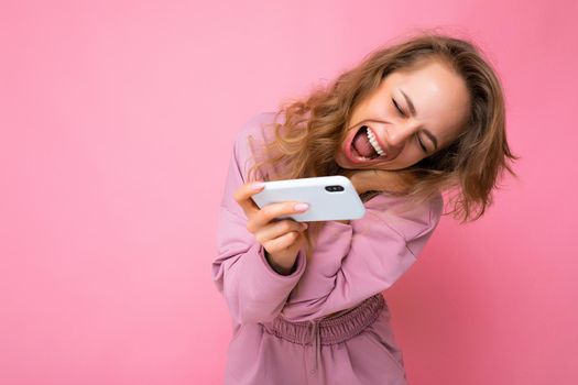 Photo of attractive crazy amazed surprised young woman wearing casual stylish clothes standing isolated over background with copy space holding and using mobile phone looking at device screen and shouting.
