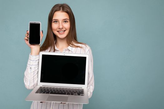 Photo of Beautiful satisfied happy young woman holding computer laptop and mobile phone looking at camera wearing shirt isolated over blue wall background. Free space, mock up