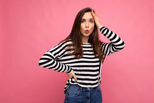 Photo of young astonished surprised beautiful brunette woman with sincere emotions wearing casual striped pullover isolated on pink background with copy space.