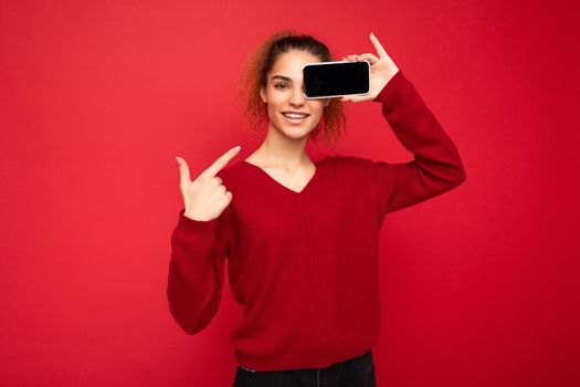 Photo of beautiful smiling young woman good looking wearing casual stylish outfit standing isolated on background with copy space holding smartphone showing phone in hand with empty screen display for mockup pointing at gadjet looking at camera.