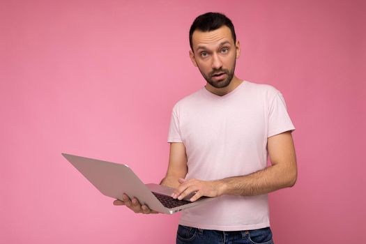 Handsome amazed and surprised man holding laptop computer looking at camera in t-shirt on isolated pink background.