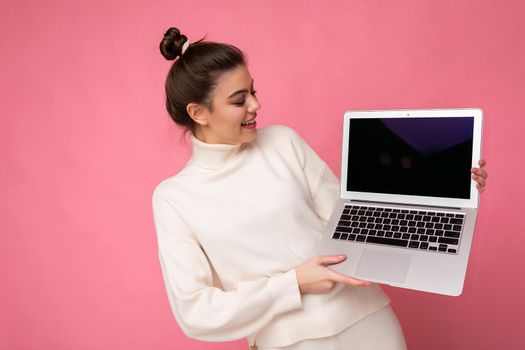 Photo of beautiful girl with gathered brunette hair wearing white sweater holding computer laptop and looking at the netbook isolated over pink wall background.