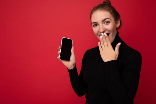 Photo of surprised attractive positive young brunette woman wearing black sweater standing isolated over red background showing mobile phone with empty screen for mockup looking at camera.