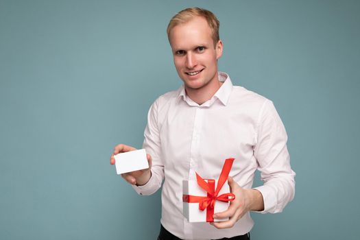 Photo shot of handsome positive smiling young blonde male person isolated over blue background wall wearing white shirt holding white gift box with red ribbon and credit card looking at camera.