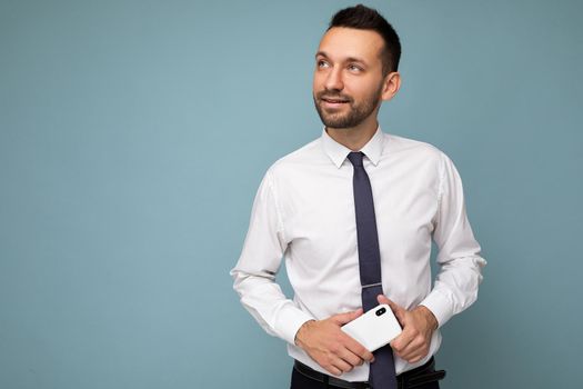 Handsome thoughtful good looking brunet unshaven man with beard wearing casual white shirt and tie isolated on blue background with empty space holding in hand mobile phone looking to the side.