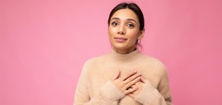 Panoramic portrait of young positive cute pretty brunette woman with sincere emotions wearing beige sweater isolated over pink background with empty space and holding hand on chest.