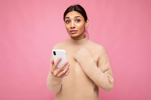 Attractive cute concerned young brunet woman wearing beige warm sweater standing isolated over pink background surfing on the internet via phone looking at camera and keeping hand on heart.