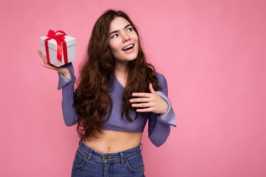 Shot of attractive positive smiling young brunette woman isolated over colourful background wall wearing everyday trendy outfit holding gift box and looking to the side. Empty space