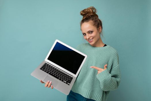 beautiful dark blond young woman with gathered curly hair looking at camera holding computer laptop with empty monitor screen with mock up and copy space wearing blue longsleeve isolated on light blue wall background.