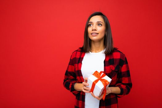 Photo shot of pretty positive young brunette woman isolated over red background wall wearing white casual t-shirt and red and black shirt holding white gift box with red ribbon and looking up. Copy space