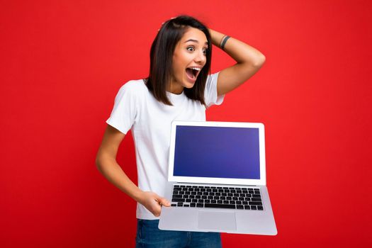 Surpriside amazed Beautiful happy overjoyed young lady with short dark brunet haircut holding computer laptop looking to the side wearing white t-shirt and jeans isolated over red wall background. Copy space for advertisement