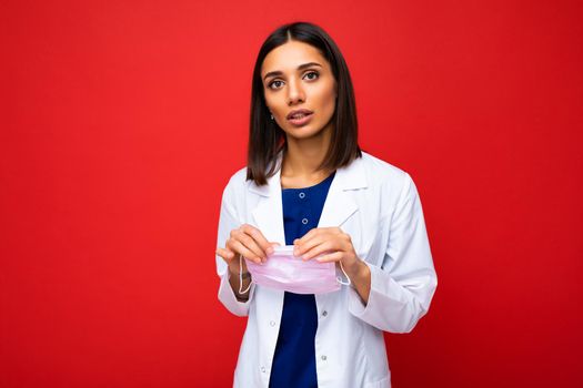 Young Woman holding virus protective mask on face against coronavirus and white medical coat isolated on red background. Free space