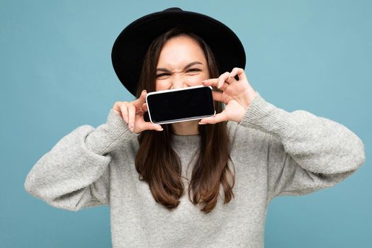 Look at this cell phone! Pleased happy woman blinks eyes, with blank screen, shows modern device, isolated over blue background. Technology concept