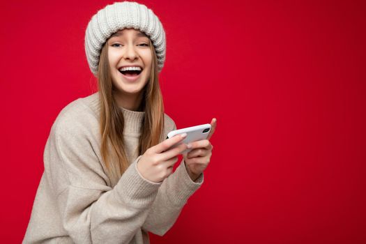 Sedi-profile photo of Beautiful crazy overjoyed young blonde woman wearing casual beige sweater and beige hat isolated over red background holding in hand and using mobile phone playing online games looking at camera. copy space