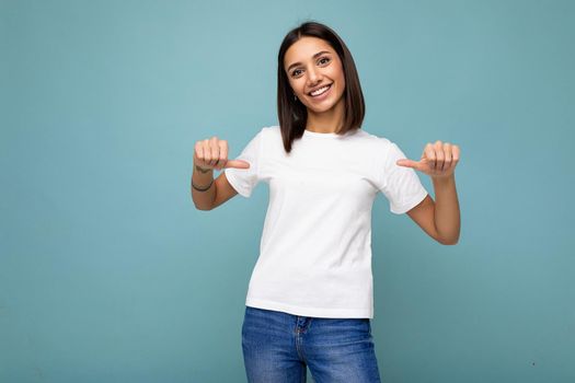 Portrait of positive happy smiling young pretty cute brunette woman with sincere emotions wearing casual white t-shirt for mockup isolated over blue background with copy space and pointing at empty space.